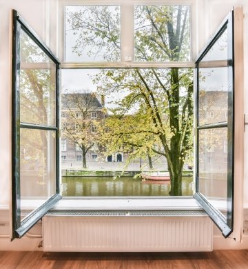 Wide-open casement glass windows with dark frames looking out over a scenic canal, autumn trees, and historic buildings in Edmonton